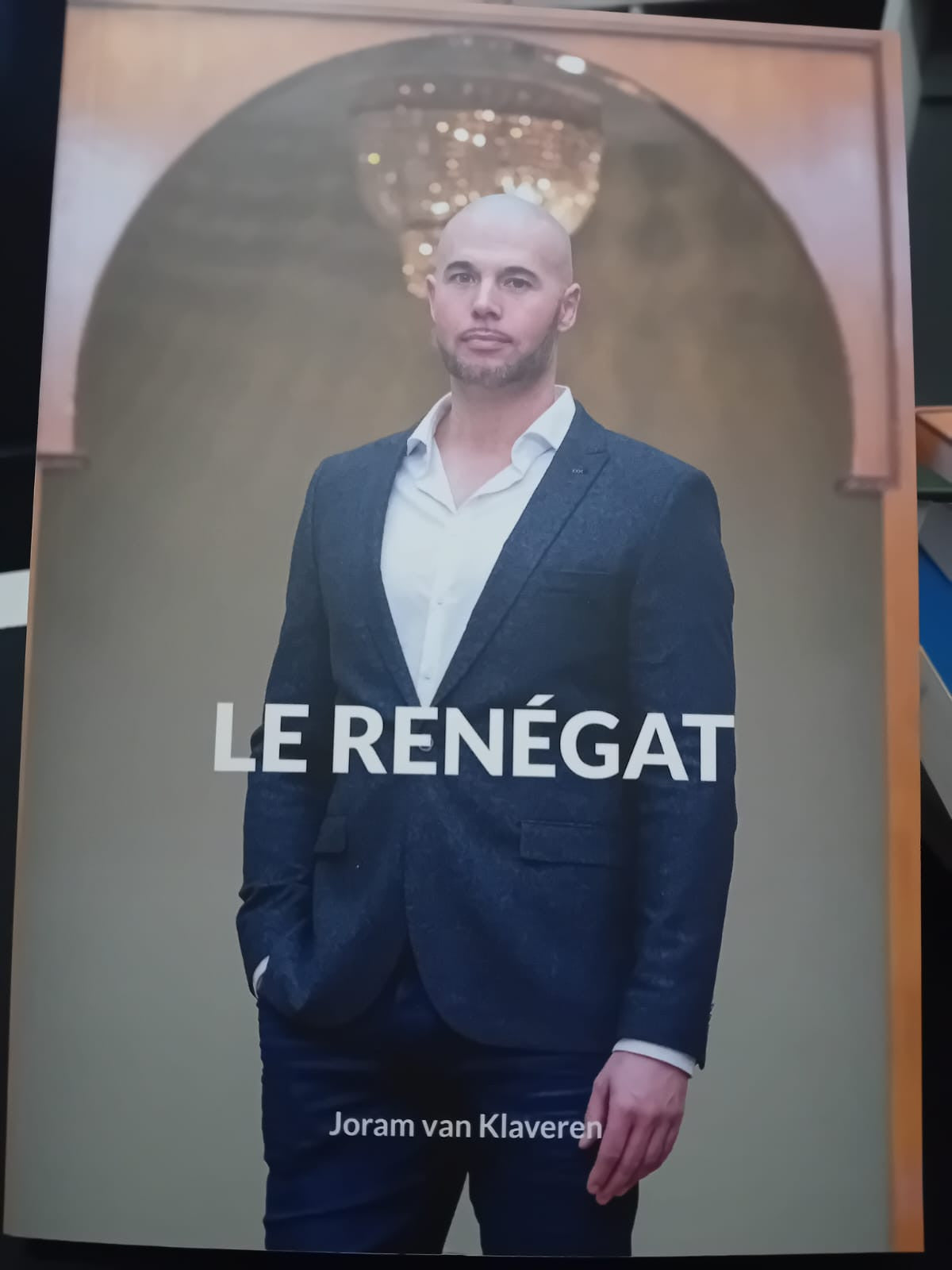 Couverture du livre Le Renégat de Joram van Klaveren, montrant l’auteur debout en costume dans une mosquée, sous un lustre, les mains dans les poches et le regard sérieux.