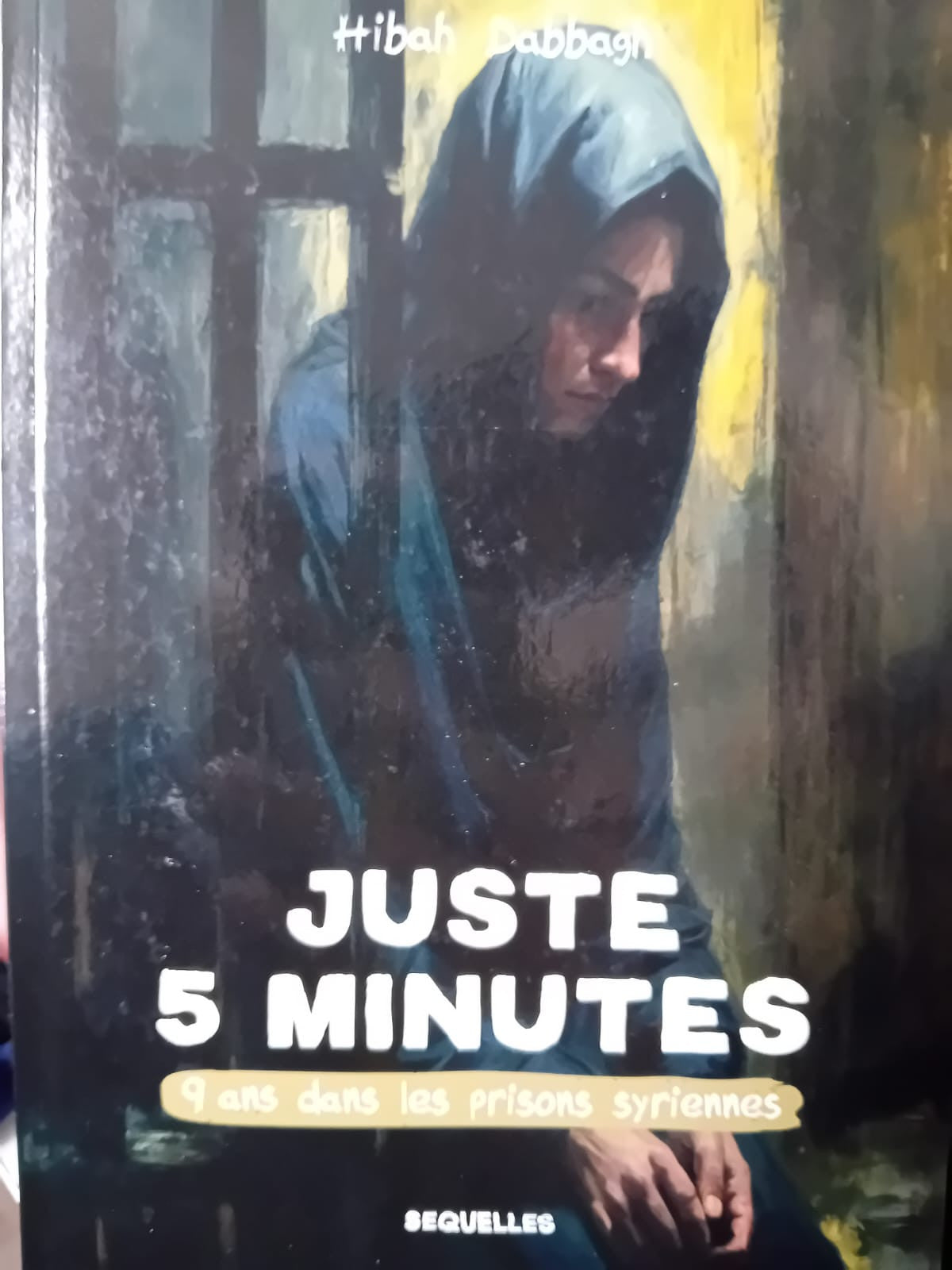 Couverture du livre Juste 5 minutes – 9 ans dans les prisons syriennes de Hibah Dabbagh, montrant une femme voilée derrière des barreaux, assise et le regard baissé, sur fond sombre et expressif.
