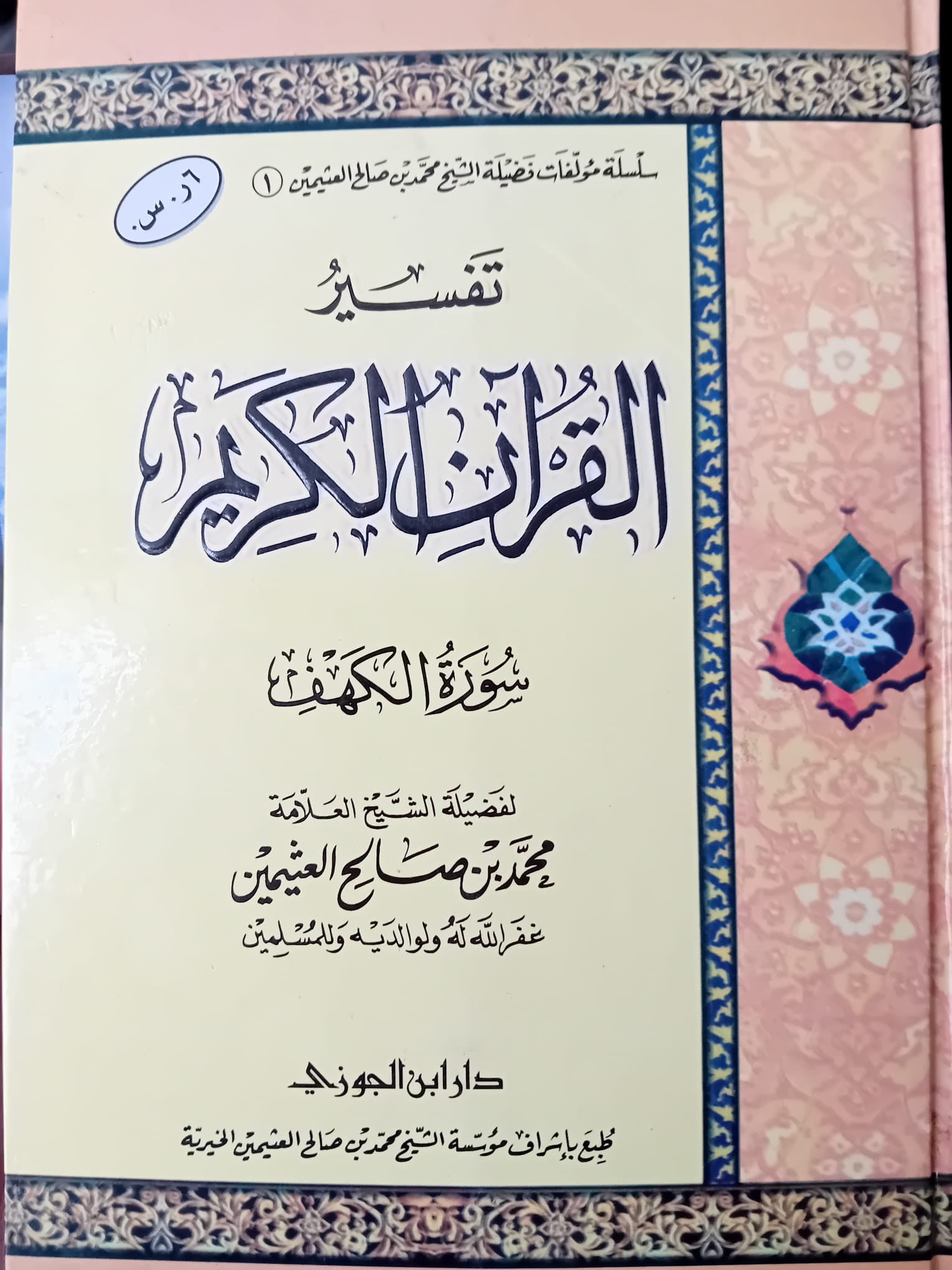 Photographie d’un livre en arabe intitulé Tafsir al-Qur'an al-Karim – Sourate al-Kahf du cheikh Muhammad ibn Salih al-‘Uthaymin, posé verticalement sur une surface claire. La couverture présente un fond crème et rose décoré de motifs islamiques et d'arabesques, avec une calligraphie soignée au centre