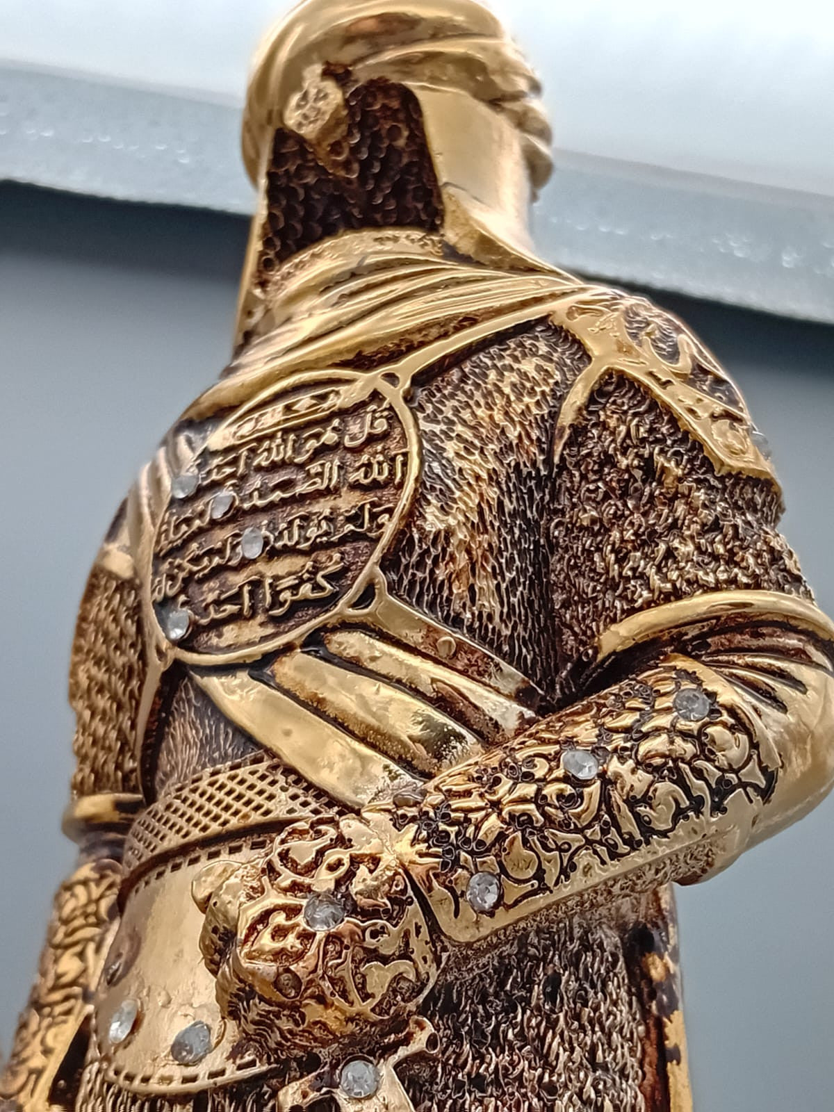 Statue dorée d’un soldat ottoman en armure, tenant un bouclier orné de calligraphie islamique, exposée sur une étagère en bois entourée de livres anciens