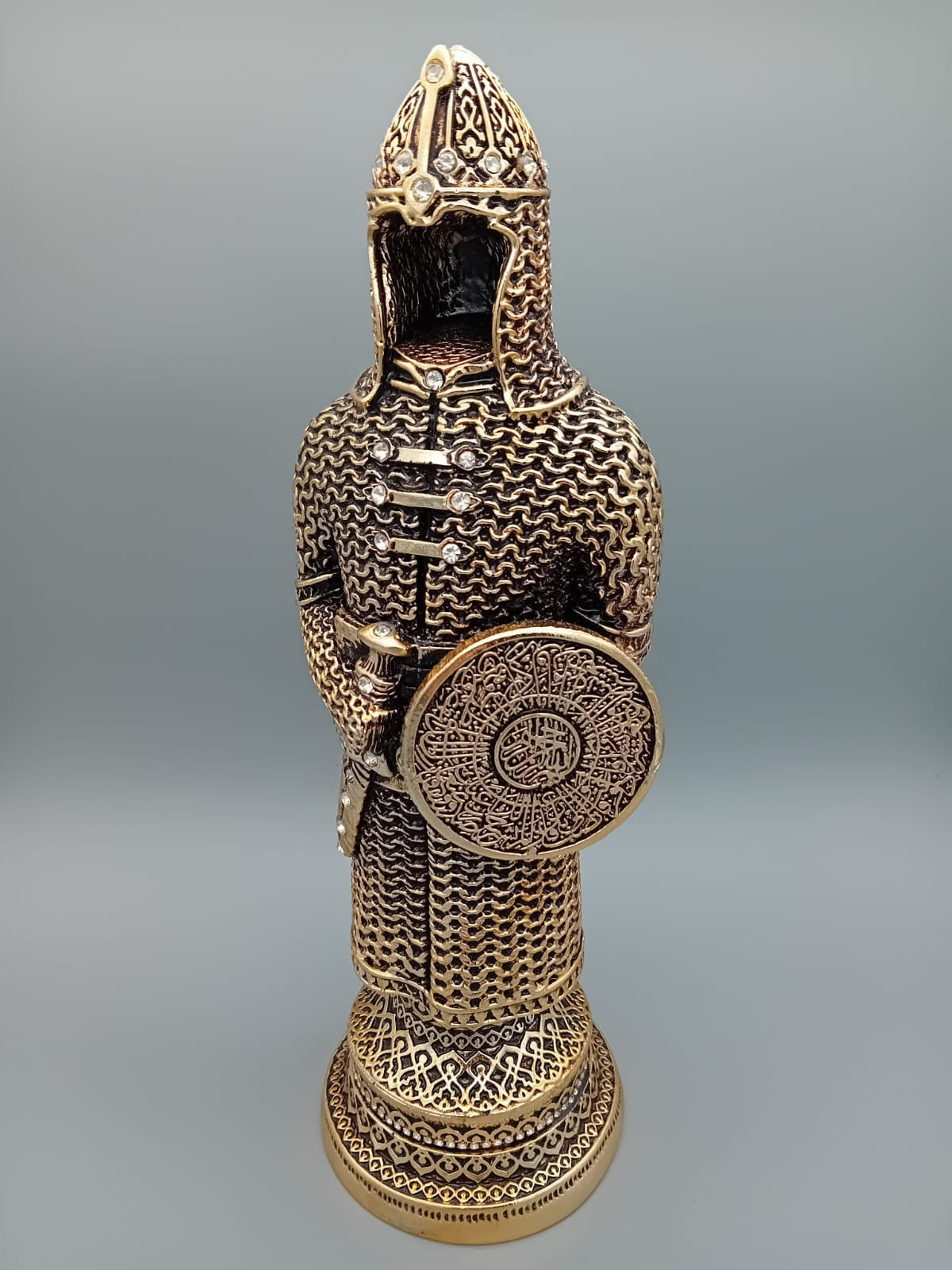 Statue décorative d’un soldat ottoman en armure, tenant un bouclier orné de calligraphies, exposée sur une étagère en bois aux côtés de livres anciens.