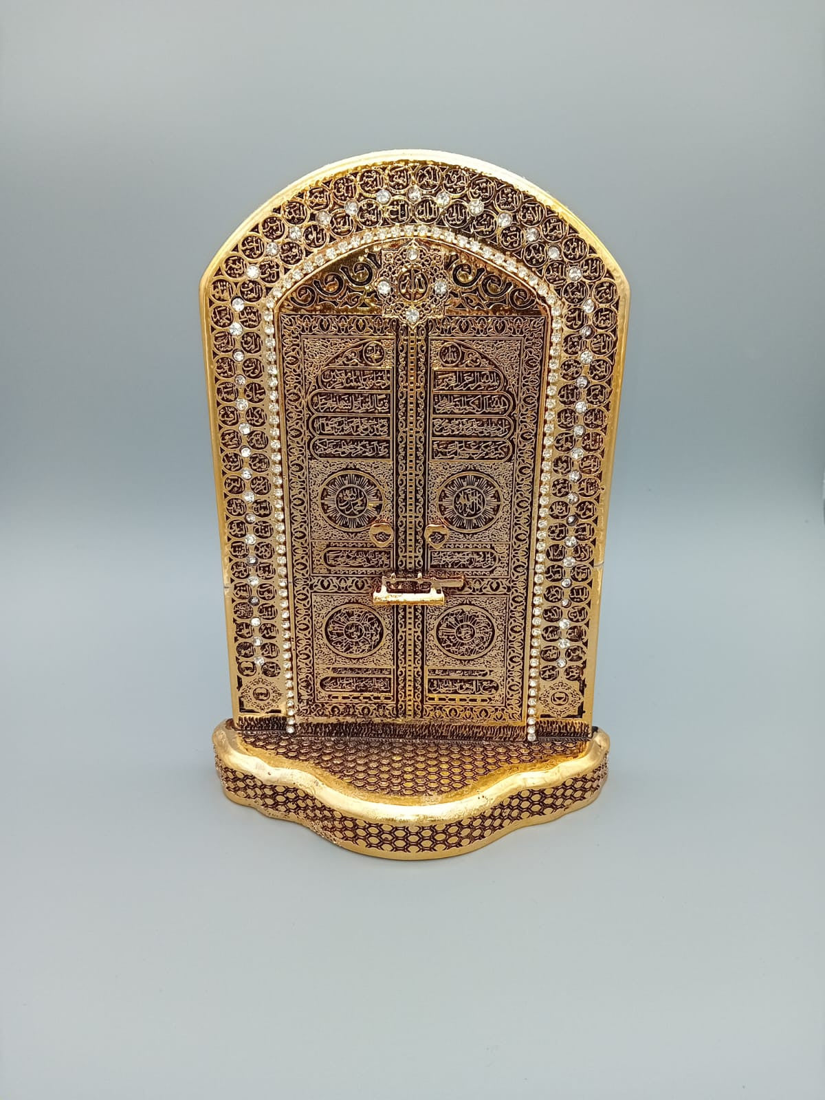 Porte Kaaba décorative dorée représentant la porte de la Kaaba, richement ornée de motifs islamiques, exposée sur une étagère en bois entre des livres anciens.