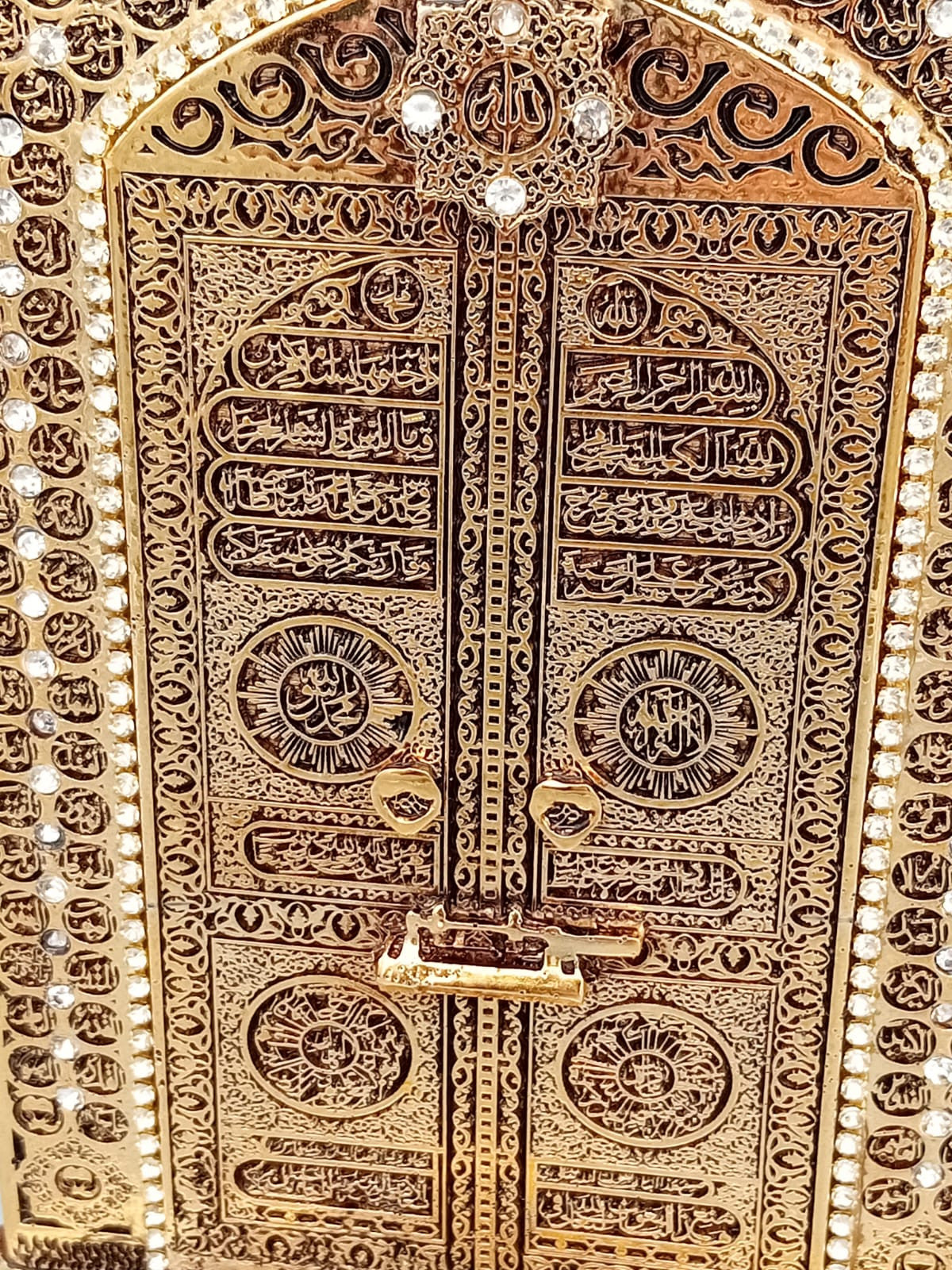 Porte Kaaba décorative dorée représentant la porte de la Kaaba, richement ornée de motifs islamiques, exposée sur une étagère en bois entre des livres anciens.