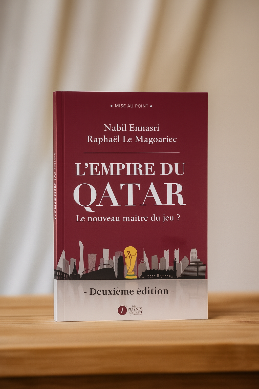 Photographie du livre L’Empire du Qatar – Le nouveau maître du jeu ? (deuxième édition) de Nabil Ennasri et Raphaël Le Magoariec, posé debout sur une table en bois clair, avec en arrière-plan un rideau blanc doux et lumineux. La couverture bordeaux du livre est mise en valeur dans une ambiance sobre et élégante.